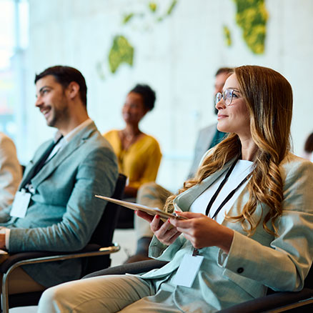 Eine Frau sitzt in einem Seminarraum, h&auml;lt ein Tablet und blickt konzentriert nach vorne. Um sie herum sitzen weitere Teilnehmende, die ebenfalls zuh&ouml;ren.