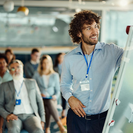 Ein Mann steht vor einem Whiteboard und zeigt auf Inhalte, w&auml;hrend mehrere Personen im Hintergrund sitzen und ihm zuh&ouml;ren.