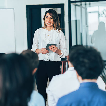 Eine Frau steht vor einer Gruppe von Zuh&ouml;renden und spricht l&auml;chelnd, w&auml;hrend sie ein Tablet in der Hand h&auml;lt. Die Teilnehmenden sitzen vor ihr und h&ouml;ren aufmerksam zu.